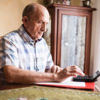 A senior man sits at a dining room table with a calculator and some bills